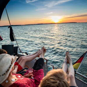 Drei Frauen an Deck einer Babro Beluga. Sie haben die Beine an der Reling hochgelegt und schauen zum Sonnenuntergang.