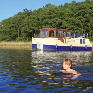 eine junge Frau schwimmt im See, im Hintergrund ankert das Hausboot