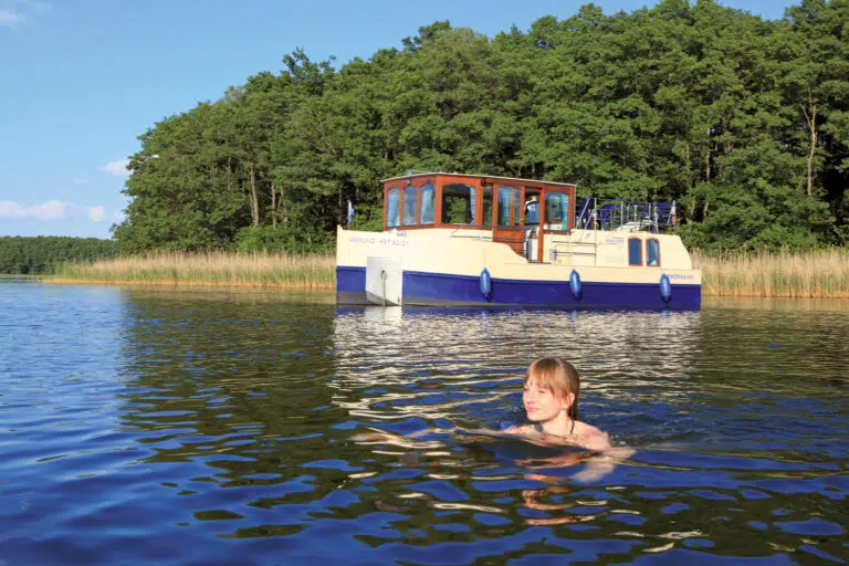 eine junge Frau schwimmt im See, im Hintergrund ankert das Hausboot