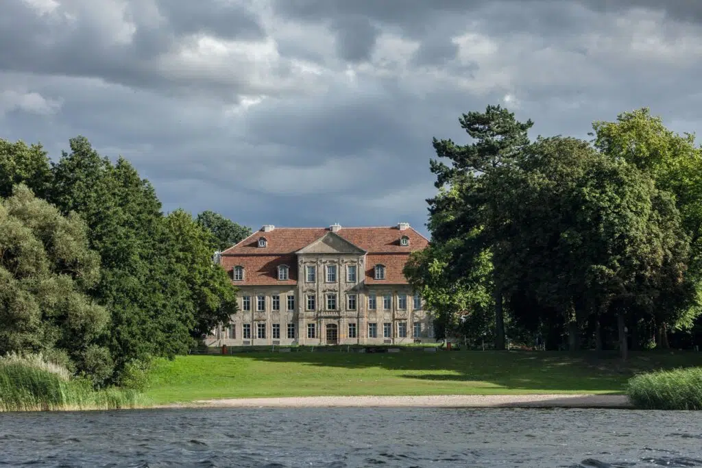 Schloss Kummerow in der Frontalansicht , leicht von Bäumen verdeckt und der See im Vordergrund