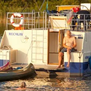 Ein Mann sitzt auf der Badeplattform einer Kormoran 1280. Neben der Kormoran sind zwei Kanus