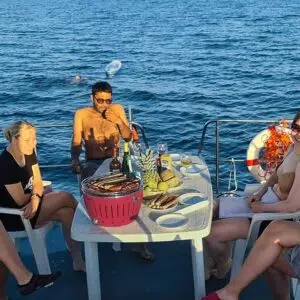 Eine Gruppe auf dem Sonnendeck der Pirate 1200. Auf dem Tisch steht ein Grill, ein Teller mit fertigen Würstchen und ein Korb mit Obst.