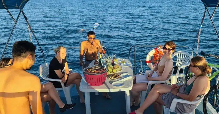 Eine Gruppe auf dem Sonnendeck der Pirate 1200. Auf dem Tisch steht ein Grill, ein Teller mit fertigen Würstchen und ein Korb mit Obst.