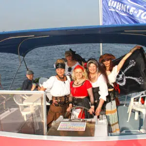 Ein paar Frauen als Piratinnen verkleidet an Bord der Pirate 1200. Eine Frau ist am Steuer, eine andere Frau hält eine Piratenflagge hinter sich hoch.