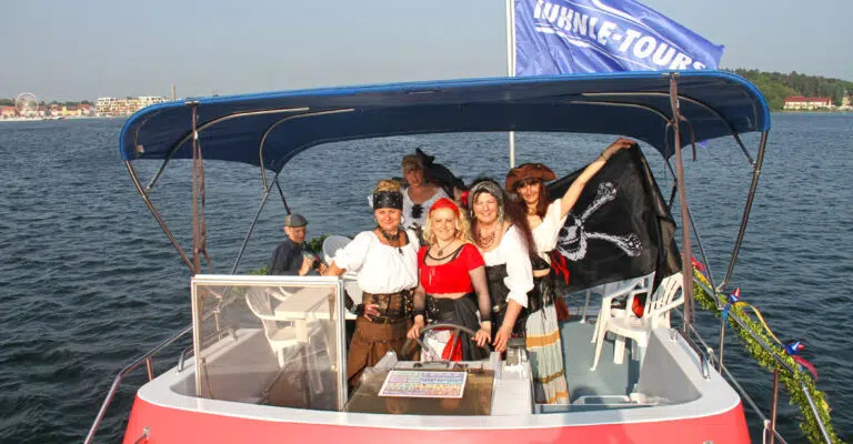 Ein paar Frauen als Piratinnen verkleidet an Bord der Pirate 1200. Eine Frau ist am Steuer, eine andere Frau hält eine Piratenflagge hinter sich hoch.