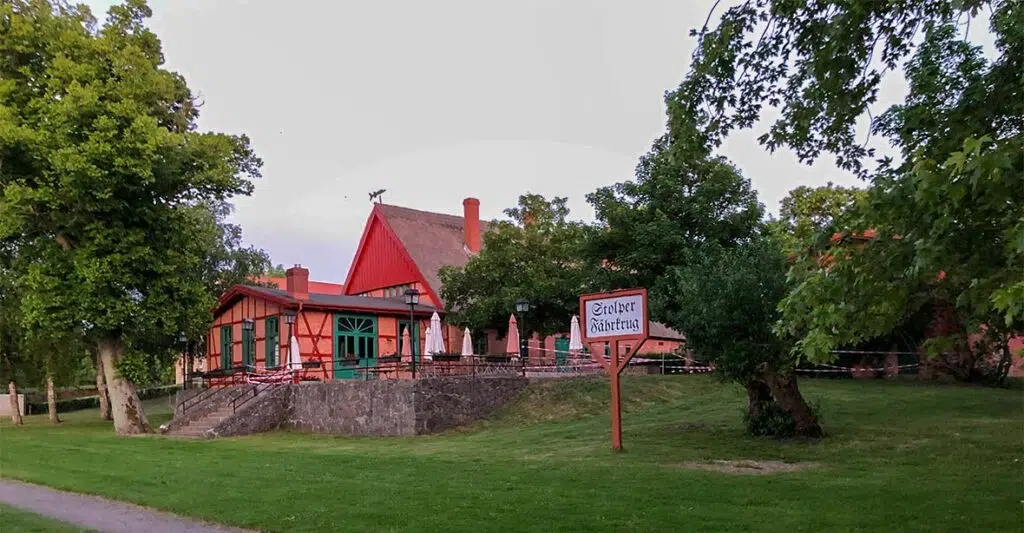 Eingang zum Stolper Fährkrug. Ein rotes Fachwerkhaus auf einer Wiese mit einigen Bäumen.