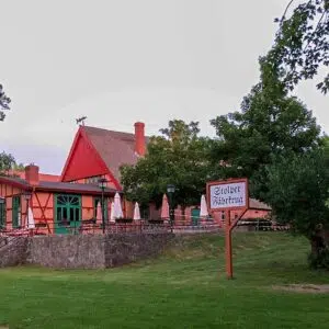 Eingang zum Stolper Fährkrug. Ein rotes Fachwerkhaus auf einer Wiese mit einigen Bäumen.