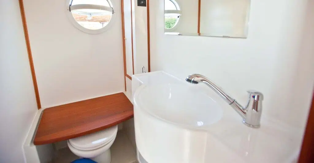 Das Badezimmer mit manueller Toilette und Waschbecken mit beweglichem Duschkopf. Die Toilette ist mit einer Platte in Holzoptik abgedeckt.