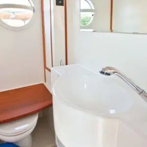 Das Badezimmer mit manueller Toilette und Waschbecken mit beweglichem Duschkopf. Die Toilette ist mit einer Platte in Holzoptik abgedeckt.
