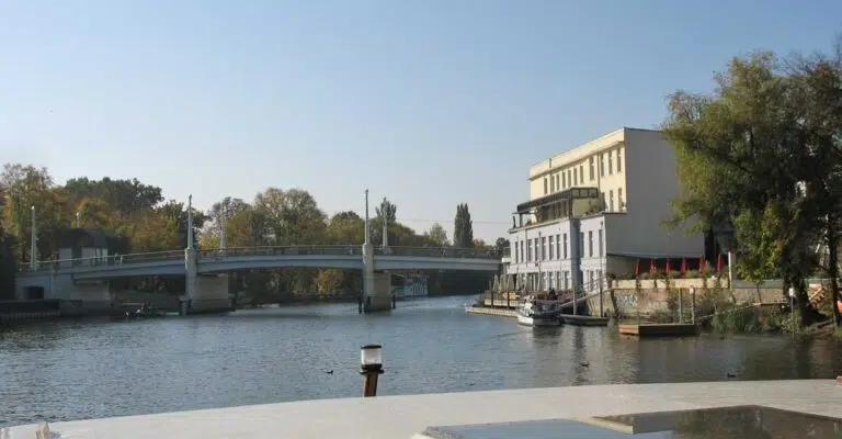 Eine Kormoran auf einem Kanal in Brandenburg mit Blick auf ein Restaurant und einer Brücke.