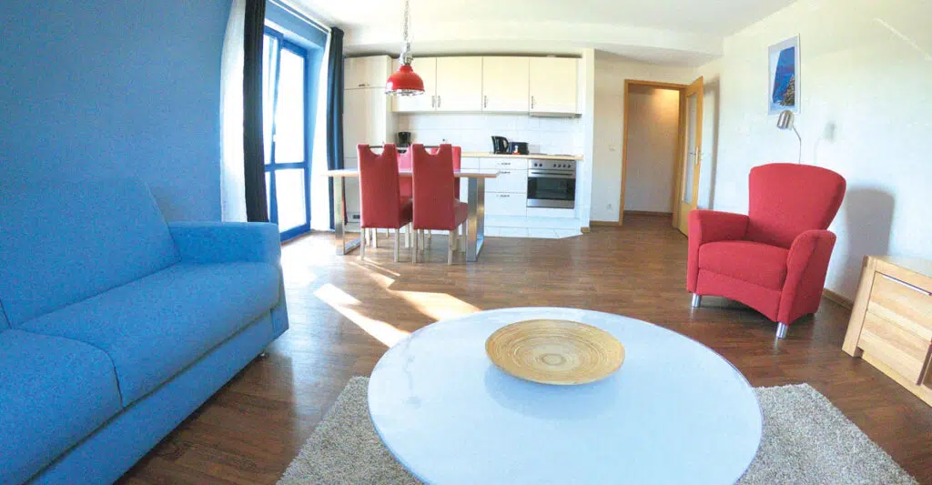 Wohnzimmer mit Couch und Sessel. Blick zur Küchenzeile mit Esstisch und Tür zum Balkon.